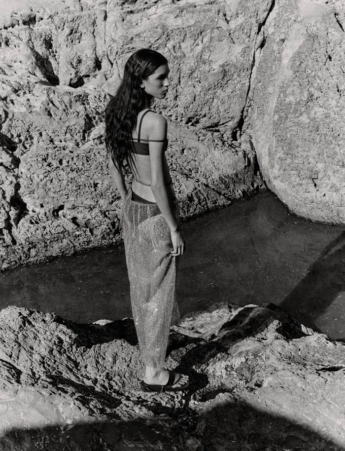 Model Jennifer Matias standing on a rugged rocky coast wearing a dark bikini top and a sheer skirt in a black and white editorial.