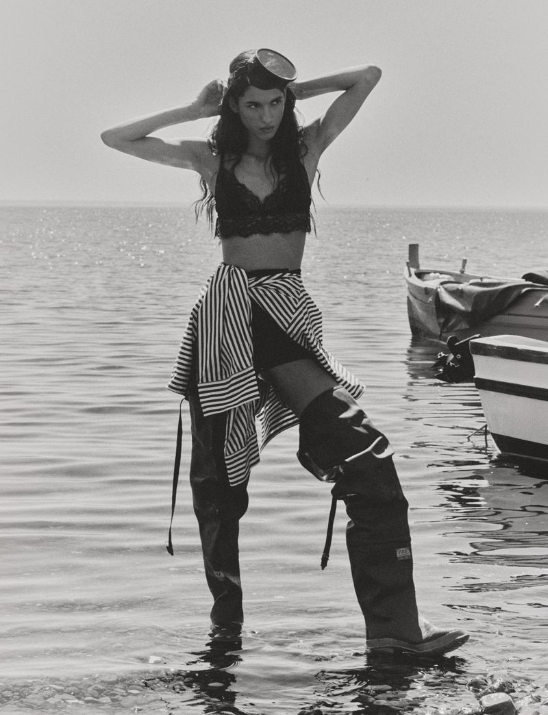 Black and white photo of Jennifer Matias standing in water wearing thigh-high rubber waders, a lace bra, and diving goggles on her head.