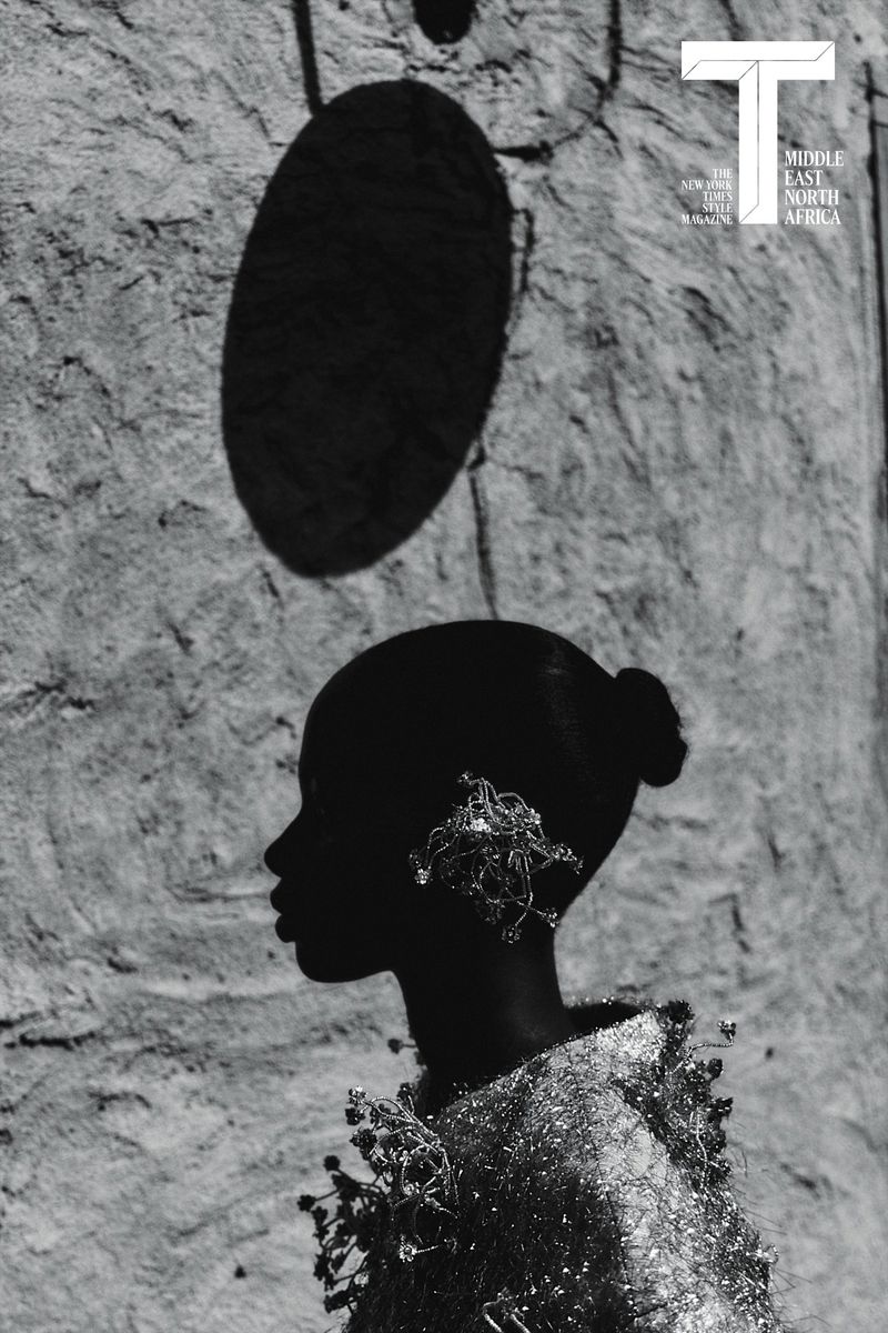 Black and white profile portrait of a woman wearing a textured garment and ornate ear jewelry against a textured stone wall.