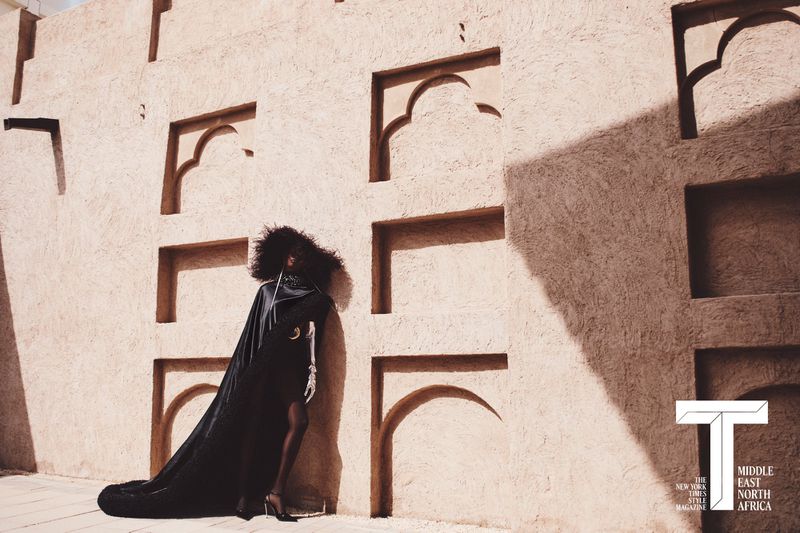 Fashion model in a voluminous afro and trailing black cloak posing against a sunlit adobe wall with decorative arched niches.