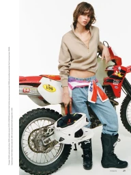 Mathilda Gvarliani posing in a beige half-zip and denim jeans beside a red dirt bike against a white background.