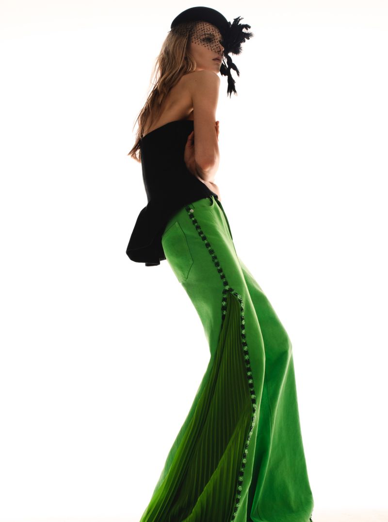 Model wearing green wide-leg trousers and a black strapless top with a netted fascinator hat against a white background.
