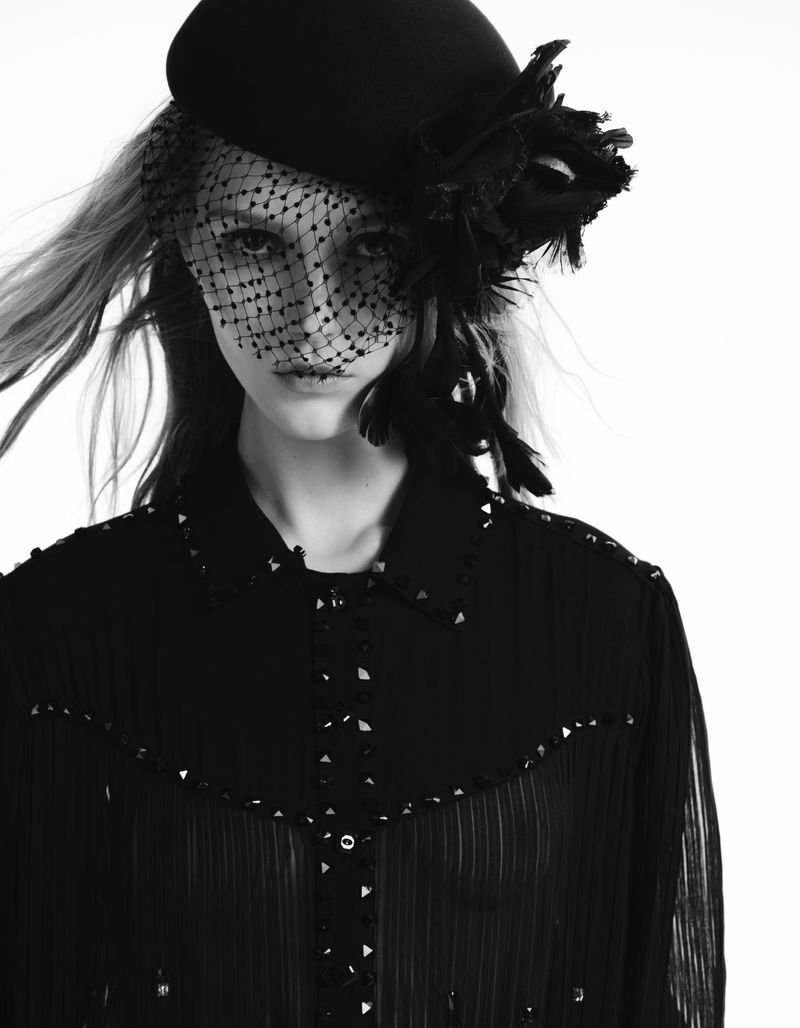Model Valeria Buldini wearing a black netted veiled beret and a studded pleated shirt against a bright white background.
