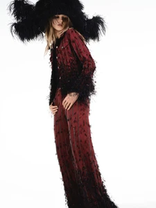 Valeria Buldini wears a burgundy fringe-trimmed jacket, matching trousers, and an oversized black feathered hat on a white background.