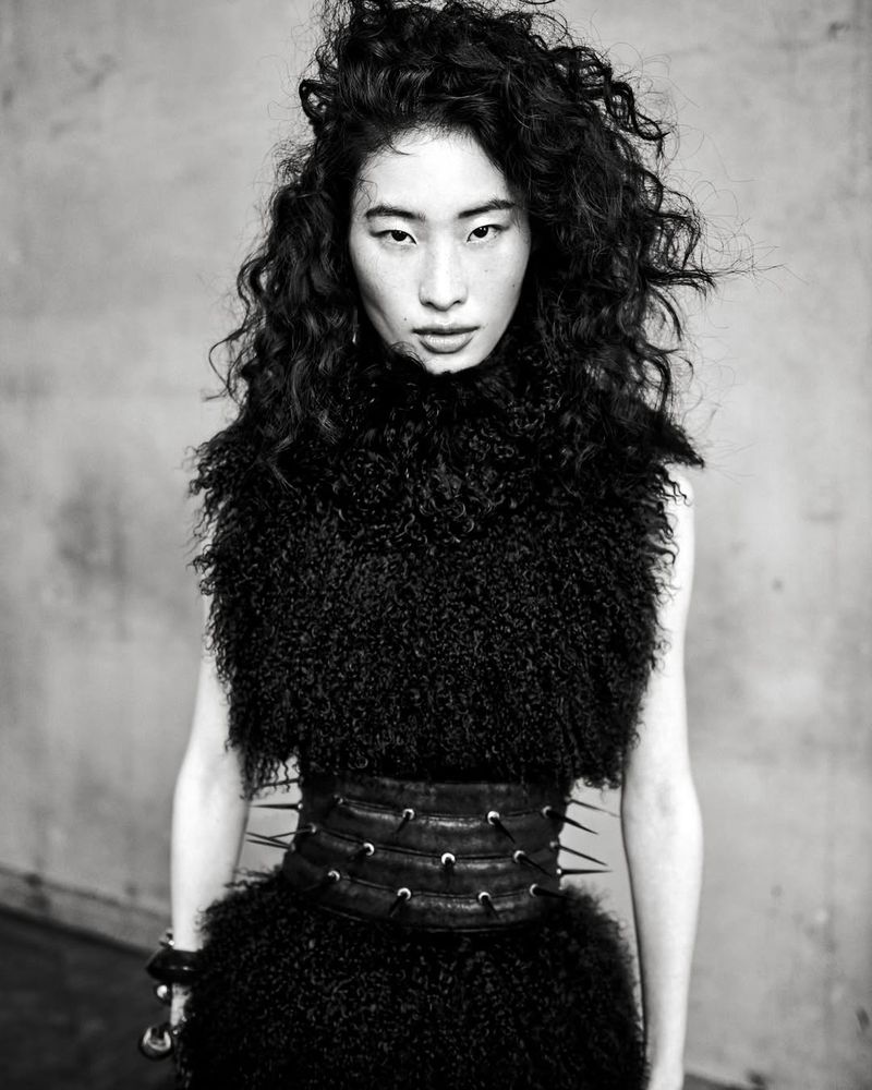 Portrait of an Asian fashion model with dark curly hair wearing a black, textured dress and spiked belt against a neutral backdrop.