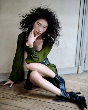 Model He Jing poses on a wooden floor wearing an olive green long-sleeve dress and black heeled boots against a neutral wall.