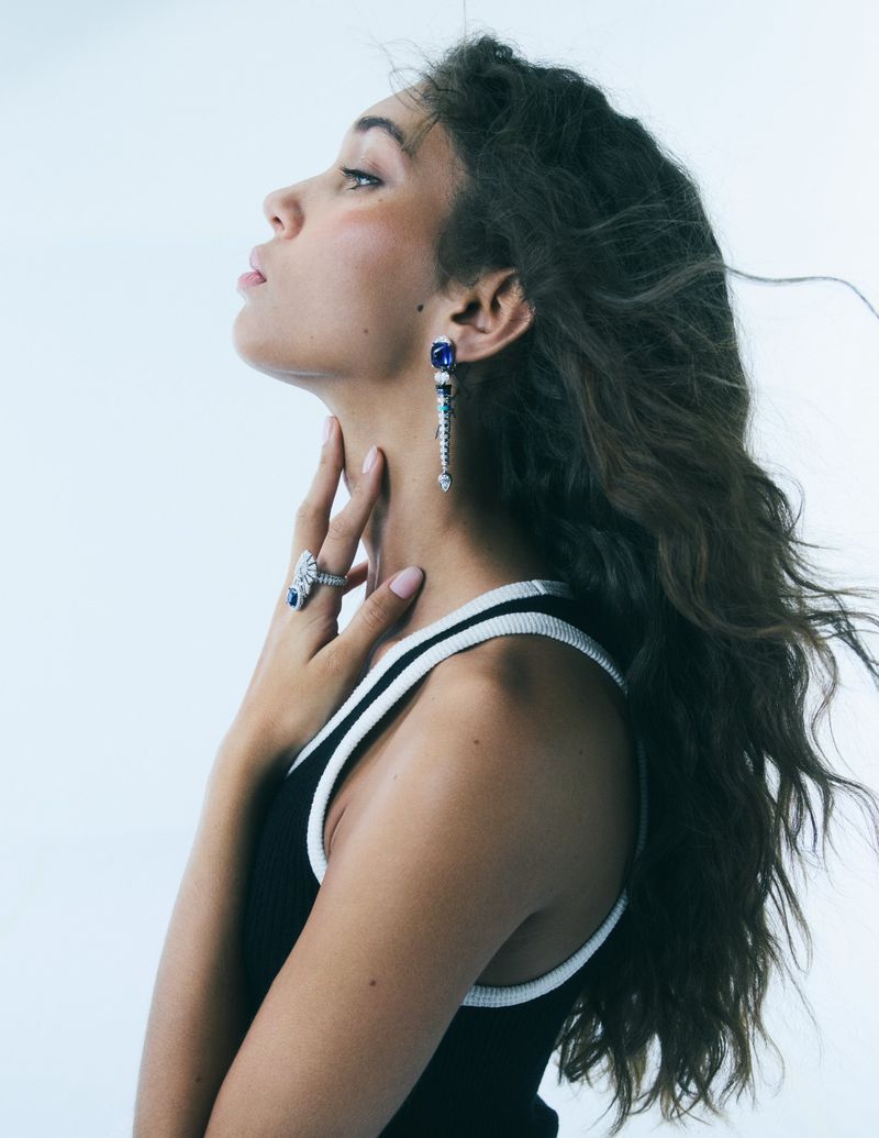 Model Latahlia Hickling in profile wearing blue gemstone drop earrings, a silver ring, and a black tank top against a white backdrop.