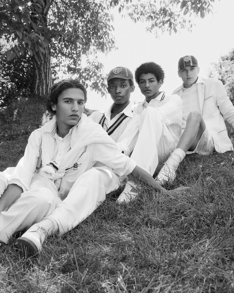 Four young male models wearing white cricket sweaters, polo shirts, and caps seated on a grassy lawn in black and white.