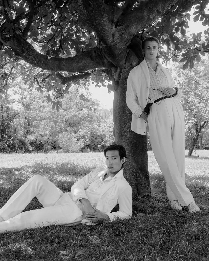 Two male models in light tailored menswear posing by a large leafy tree in a black and white fashion editorial shot.