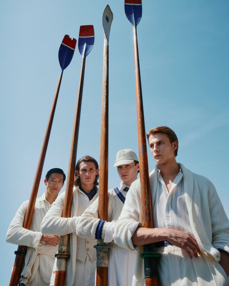 Four male models in cream knitwear holding long wooden oars against a clear blue sky background.