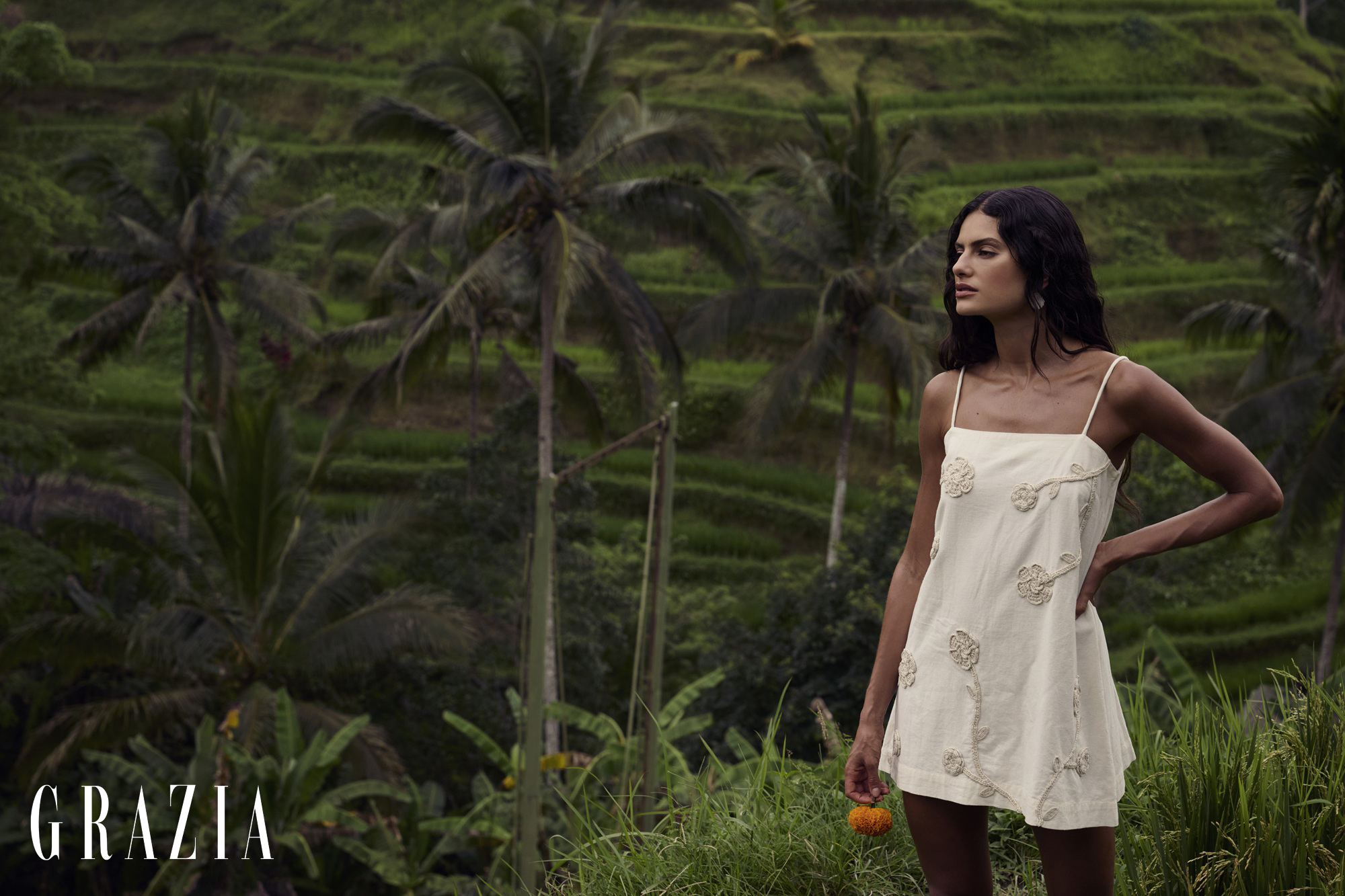 Woman in an ivory floral embroidered mini dress standing before lush green terraced hills holding a small orange flower.