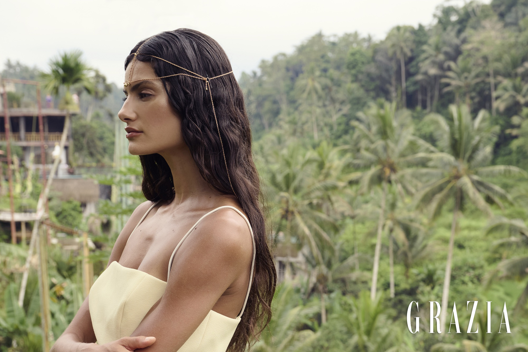 Profile portrait of a brunette woman wearing a delicate head chain and yellow dress against a lush tropical palm forest.