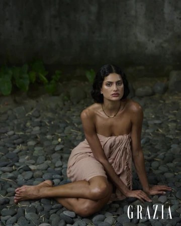 Woman seated barefoot on dark river stones wrapped in a fringed blush pink textile in an outdoor editorial portrait.