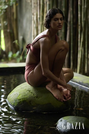 Woman in a terracotta bikini crouching on a mossy rock in a shallow pool surrounded by bamboo stalks