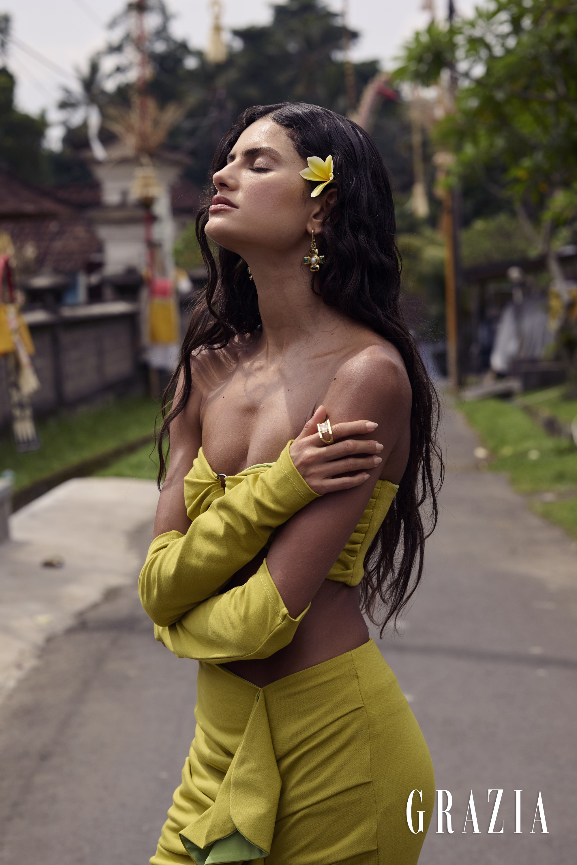 Woman with a yellow flower in her hair wearing a chartreuse strapless top, sleeves, and skirt along an outdoor paved road.