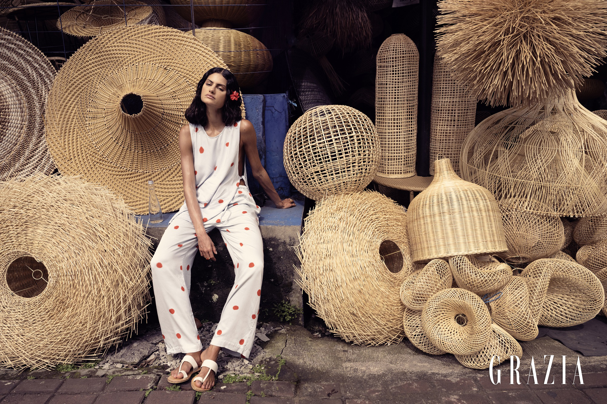 Model wearing a white polka dot jumpsuit seated amidst an array of woven rattan lampshades and sculptures for Grazia Slovenia
