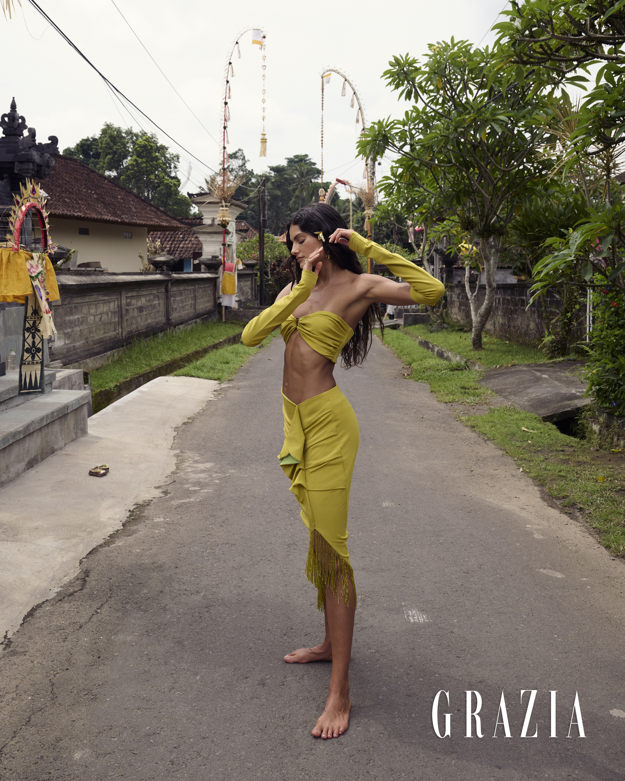 Model standing on a paved road wearing a yellow two-piece outfit with fringe details in an outdoor setting