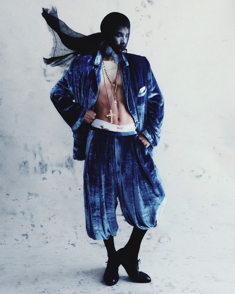 Adult male in a blue velvet jacket and pants with beaded necklaces and a dark head covering posing in front of a textured white wall.