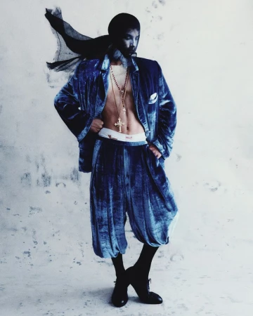 Adult male in a blue velvet jacket and pants with beaded necklaces and a dark head covering posing in front of a textured white wall.