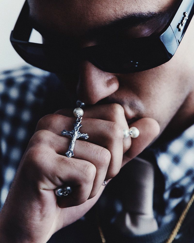 Portrait of an adult with sunglasses and silver cross necklace, hand covering the mouth, wearing a plaid shirt indoors.