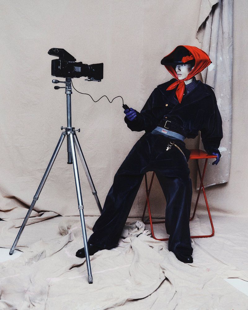 Retro camera on tripod next to a stylish person in a red scarf and dark blue suit posing against a neutral studio backdrop.
