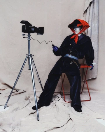 Retro camera on tripod next to a stylish person in a red scarf and dark blue suit posing against a neutral studio backdrop.