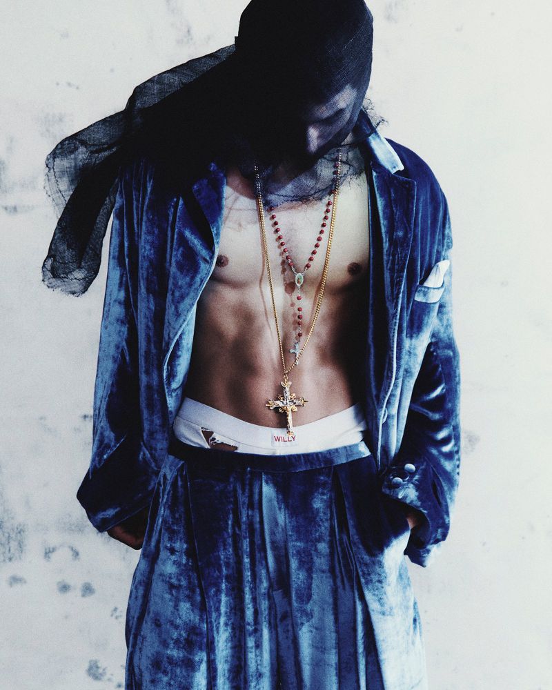 Adult male in a blue velvet suit with gold necklaces, wearing a black head covering, standing against a white painted brick wall.