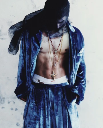 Adult male in a blue velvet suit with gold necklaces, wearing a black head covering, standing against a white painted brick wall.