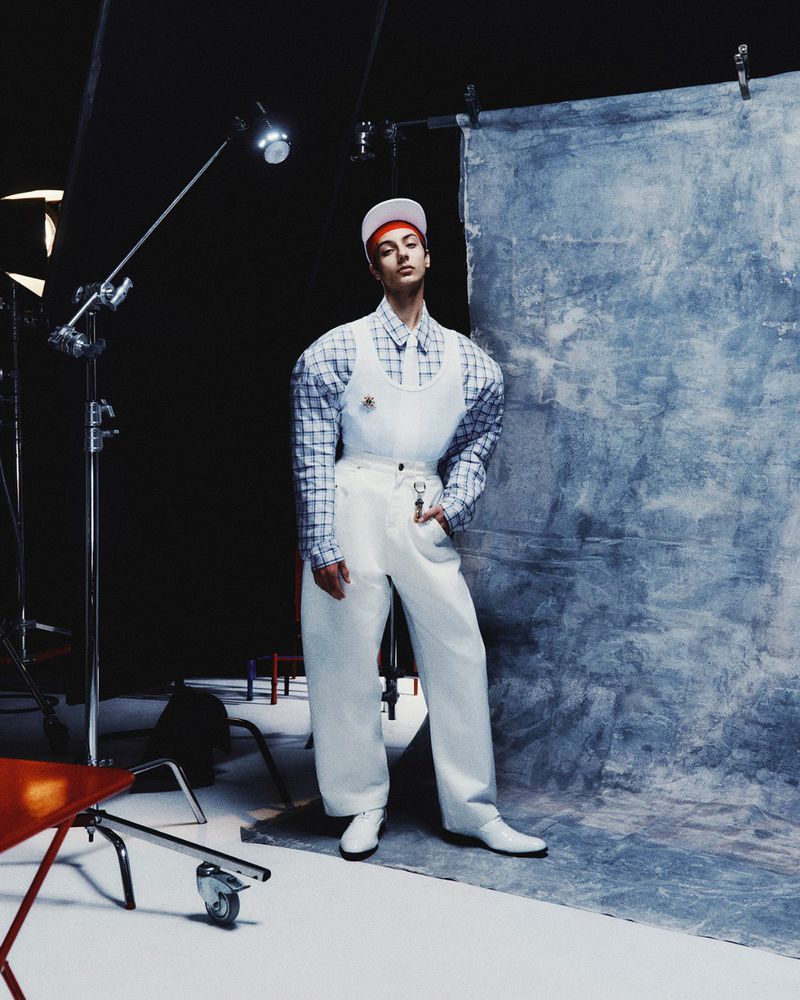A young adult poses in a white outfit with a red cap in a studio setting, illuminated by studio lights, next to a grey painted canvas.