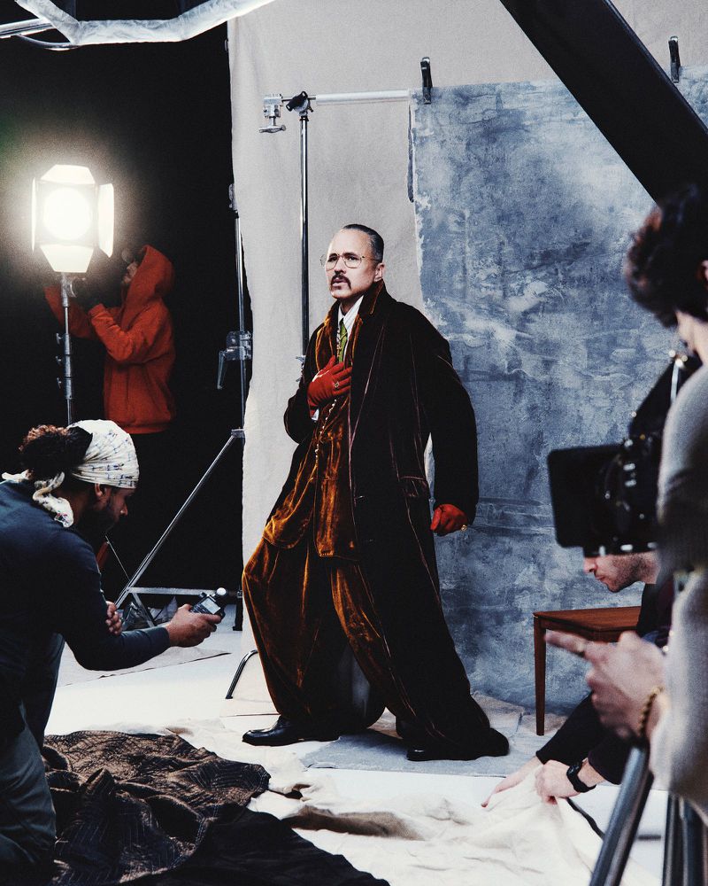 Man in velvet suit posing in a photo studio with lighting equipment and photographers visible, indicating a fashion or editorial shoot.