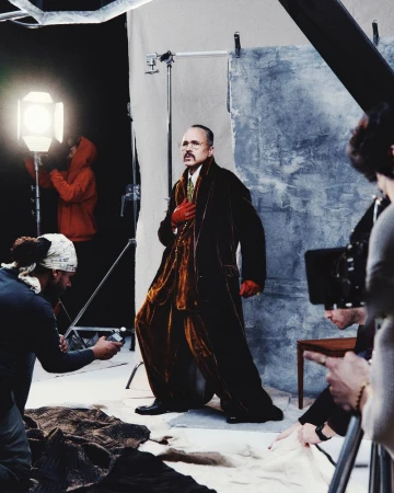 Man in velvet suit posing in a photo studio with lighting equipment and photographers visible, indicating a fashion or editorial shoot.