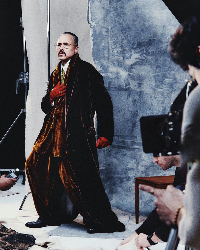 A man poses in a brown velvet suit with red gloves in front of a mottled backdrop, captured during a fashion photography studio session.