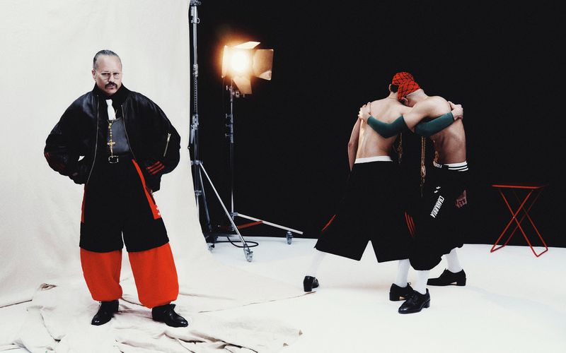 Fashion photo shoot with a man in a clerical-inspired outfit standing by two masked shirtless men embracing in a studio setting.