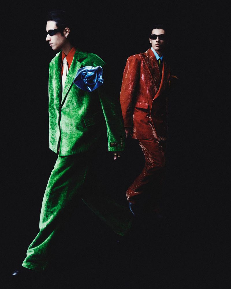 Two men in vivid velvet suits and sunglasses walk against a dark backdrop in a studio setting, showcasing unique fashion and style.