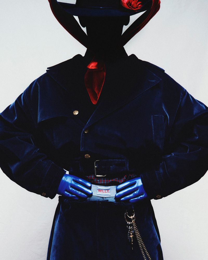 Fashionable person wearing a navy coat, hat, and gloves, posing against a white background. Stylish outfit accessorized with a belt and chain.