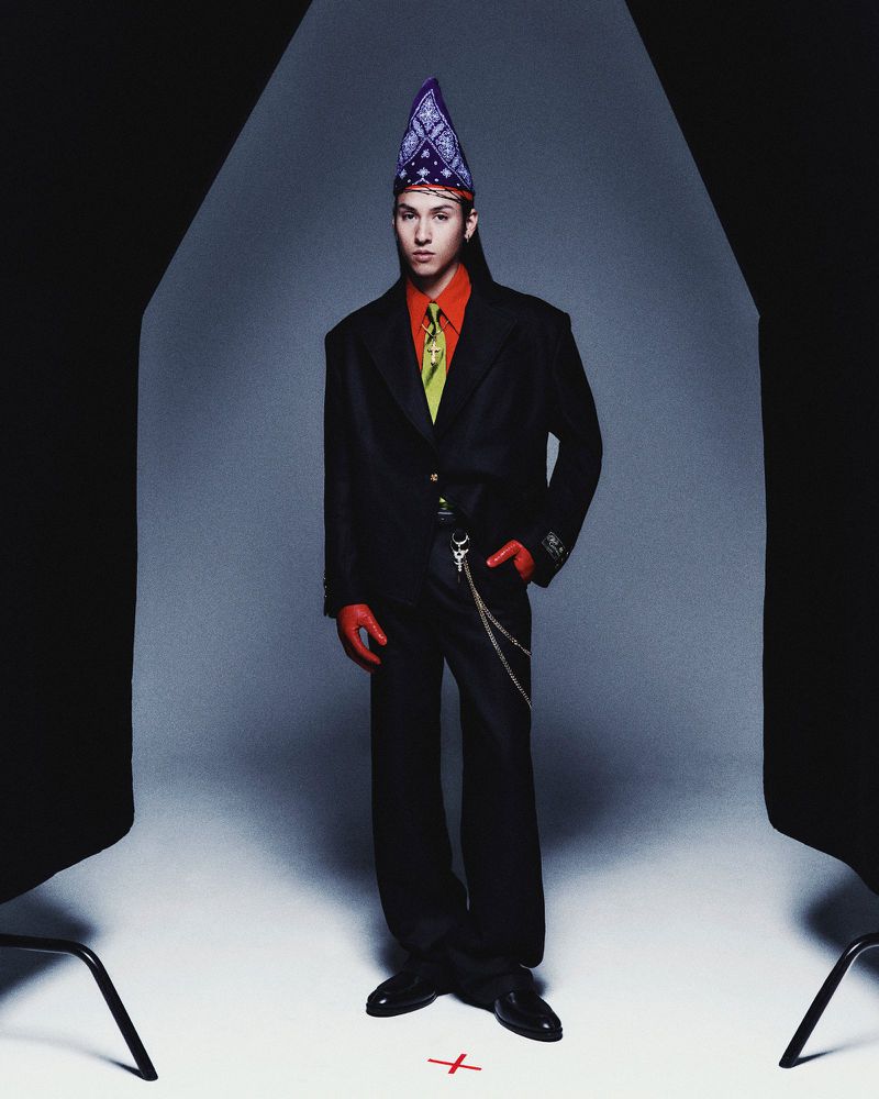 Full-length studio shot of an adult male in a black suit, red shirt and gloves, wearing a bandana hat and standing in front of a grey background.