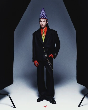Full-length studio shot of an adult male in a black suit, red shirt and gloves, wearing a bandana hat and standing in front of a grey background.