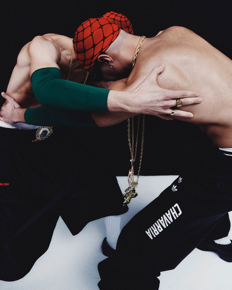 Two shirtless men in a photo studio, wearing black pants and gold jewelry, are leaning on each other against a black background.