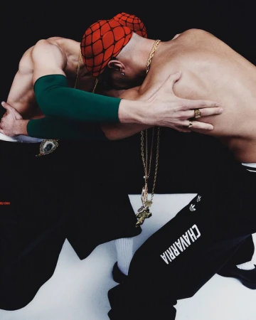 Two shirtless men in a photo studio, wearing black pants and gold jewelry, are leaning on each other against a black background.