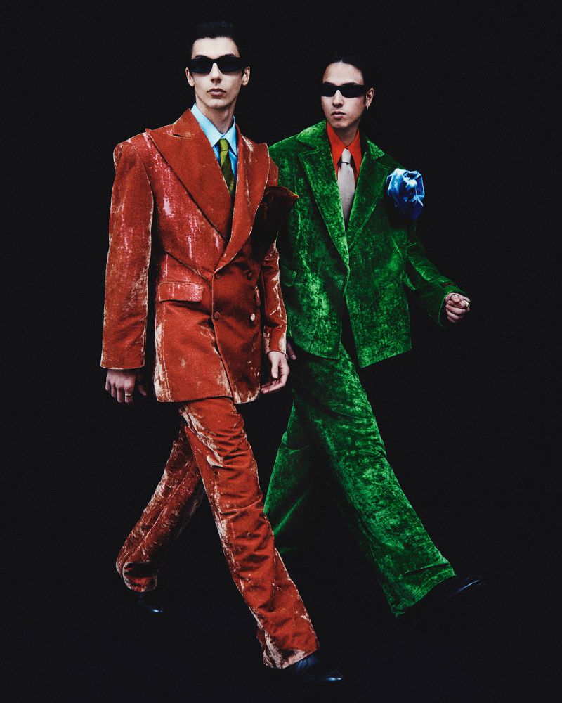 Two fashion models in vibrant velvet suits and sunglasses walk against a dark background, showcasing retro-inspired style.