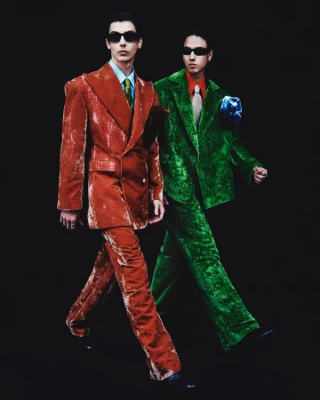 Two fashion models in vibrant velvet suits and sunglasses walk against a dark background, showcasing retro-inspired style.