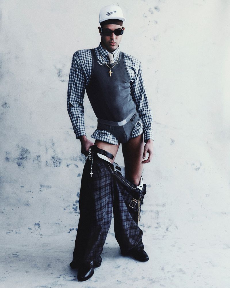 A man in dress shirt and underwear stands in front of a grey background with his pants pulled down around his ankles.