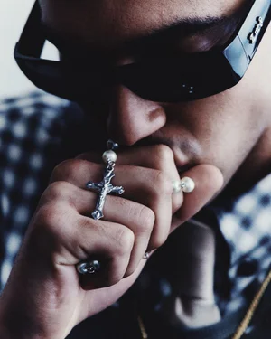 Close-up of a man wearing black sunglasses holding a pearl rosary with a silver crucifix to his mouth.