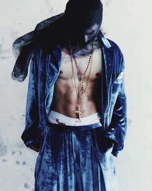 Model wearing an open blue crushed velvet suit and black mesh head covering with cross necklaces against a textured backdrop.