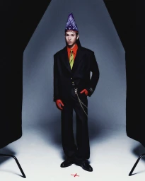 Model in a black suit, red shirt, and patterned blue bandana hat standing against a grey studio backdrop.