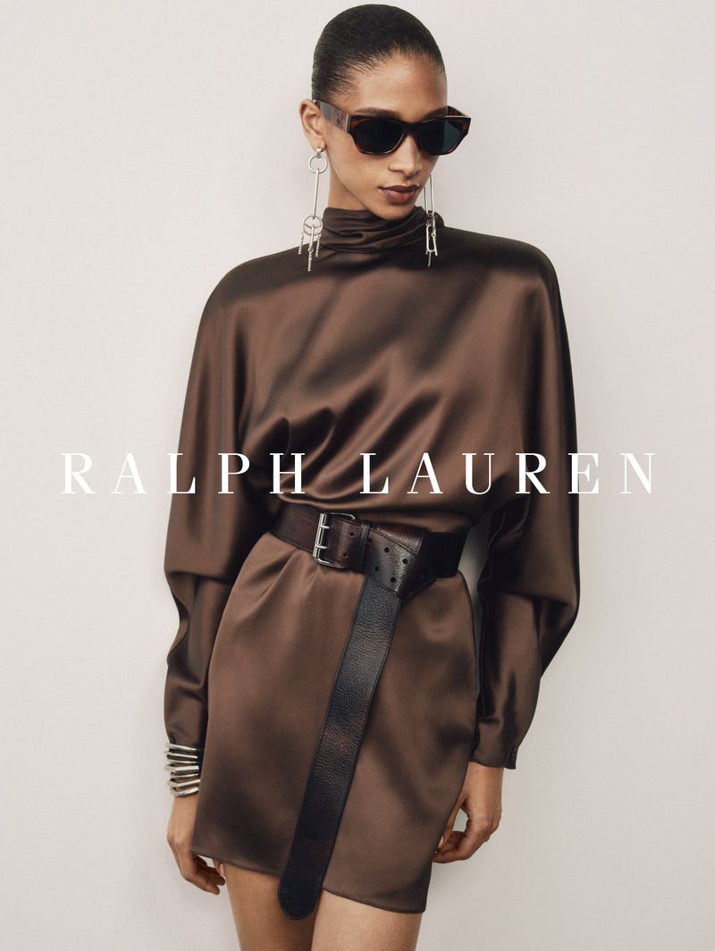 Model wearing a brown satin high-neck mini dress, dark sunglasses, and a wide leather belt against a pale studio backdrop.