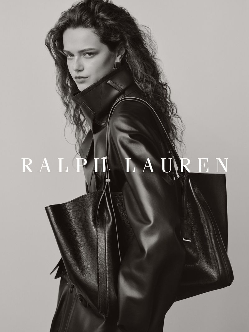 Fashion model wearing a black leather coat and carrying an oversized black leather tote bag against a plain studio backdrop.