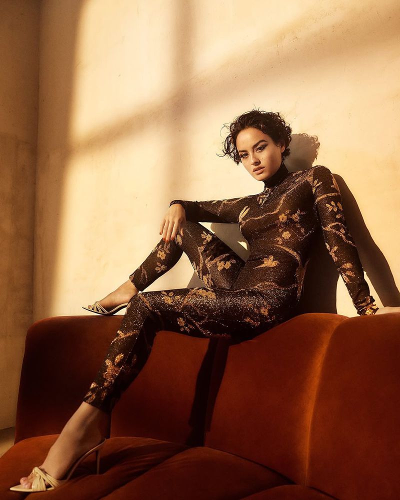 Brunette woman in floral jumpsuit poses on a rust-colored velvet couch in a warm-toned indoor studio with light and shadow play.
