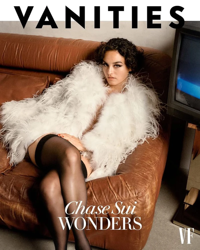 Chase Sui Wonders seated on a brown leather sofa wearing a white feathered coat and black stockings for Vanity Fair U.S.