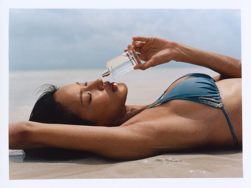 Model Hyunjoo Hwang lies on a sandy beach holding a Salt & Stone fragrance bottle near her face while wearing a teal bikini.